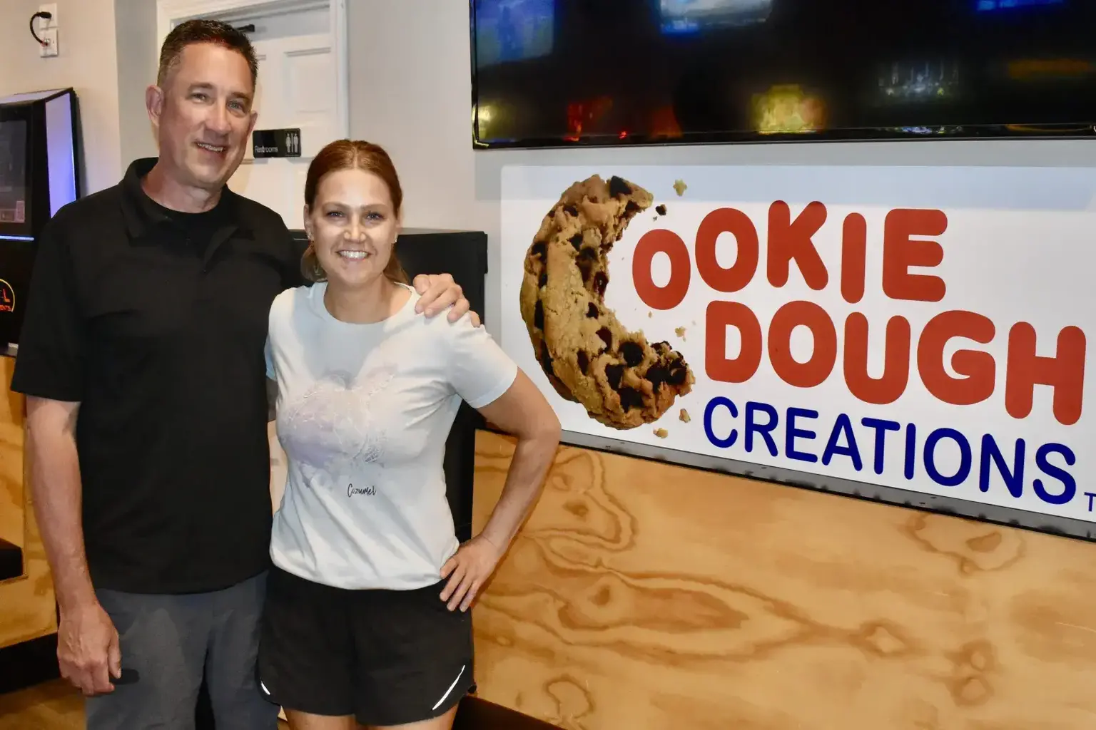 Jeff and Nikki Schultz standing together in front of the Cookie Dough Creations sign inside the shop