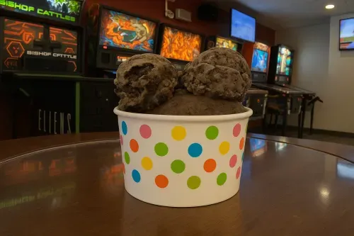 A polka-dot cup of chocolate ice cream scoops on a table in front of the lit arcade cabinets