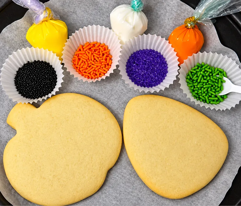 Two plain pumpkin and candy corn sugar cookies with bags of colored icing and cups of sprinkles