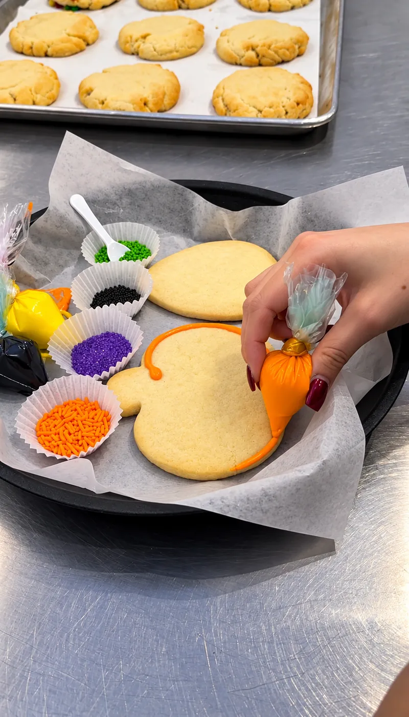 Piping orange icing onto a pumpkin-shaped sugar cookie, with a tray of fresh cookies behind it
