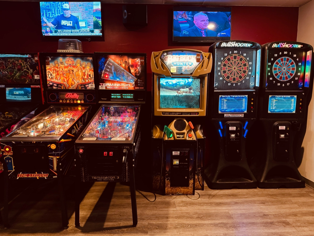 Pinball machines, an arcade cabinet and two electronic dart machines along the game room wall