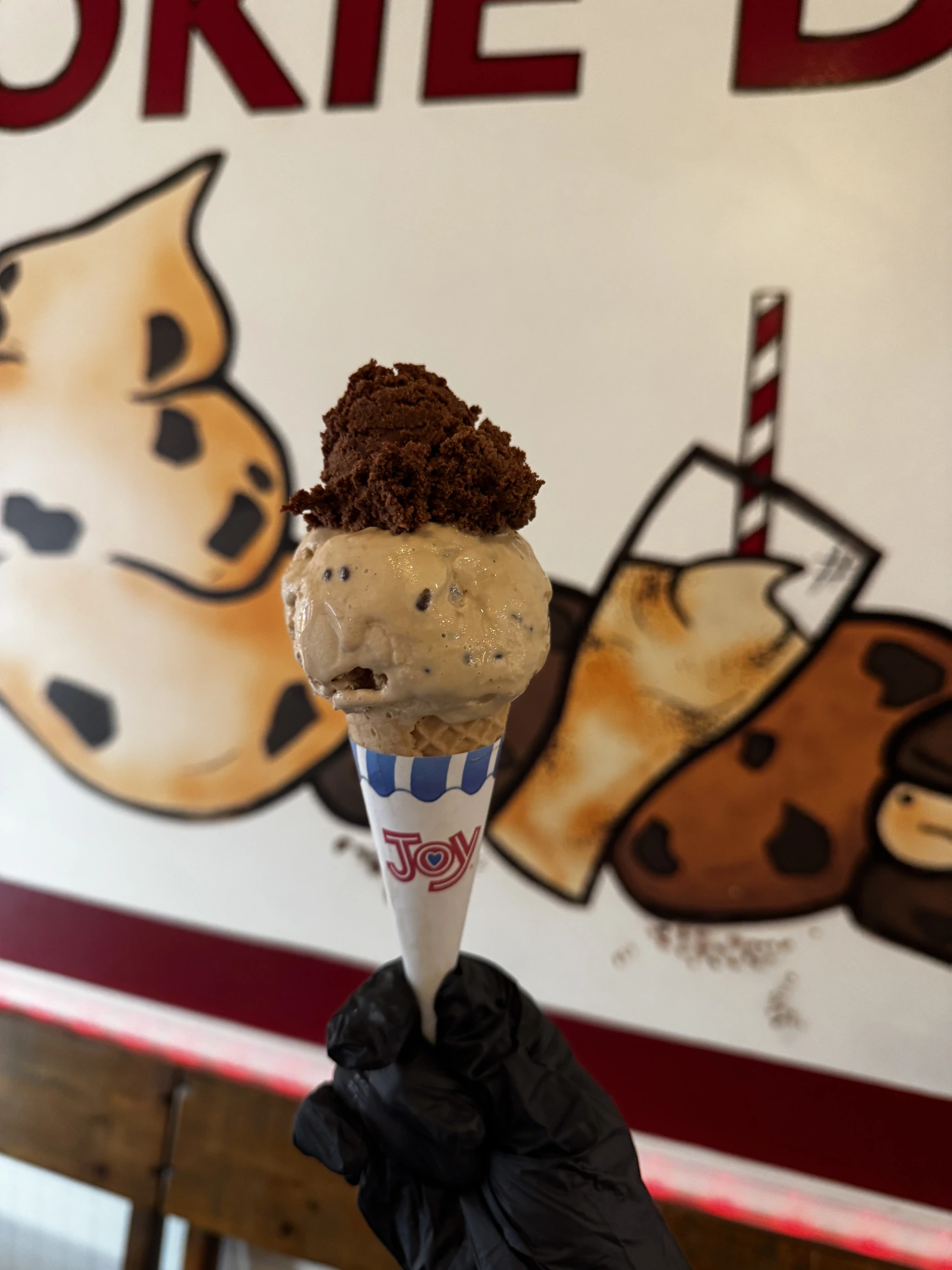 A hand-scooped waffle cone held up in front of the Cookie Dough Creations wall mural