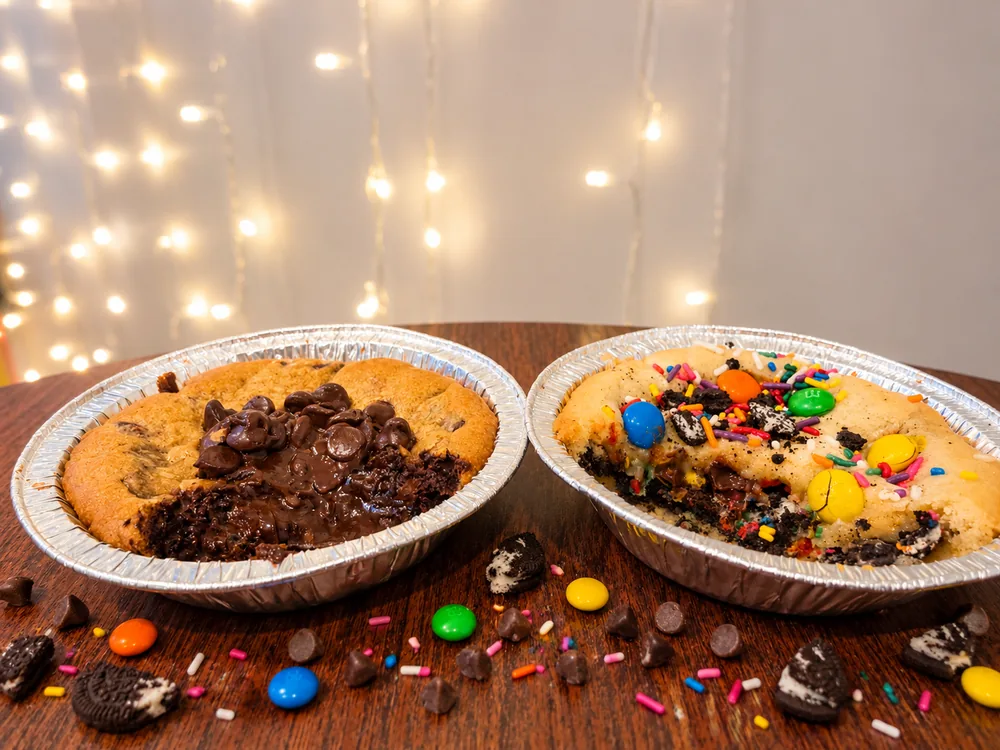 Two baked cookie pies side by side in foil tins on a wood table, one piled with chocolate chips over a fudge brownie center and one topped with M&M's, crushed Oreo cookies and rainbow sprinkles, with candy and sprinkles scattered in front and string lights behind